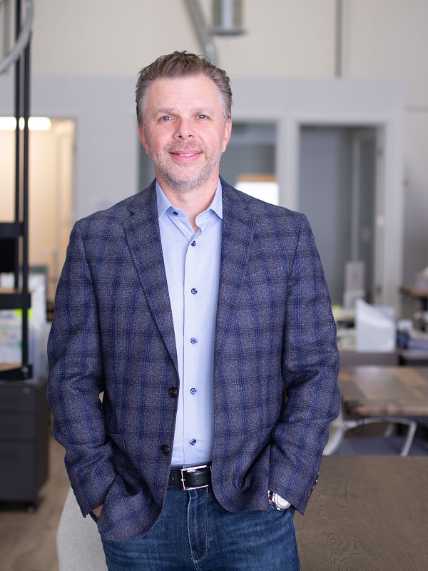 ShiraTronics CEO Rob Binney standing for a photo dressed in jeans, a blue button shirt, and a gray and blue patterned blazer with his hands in his pockets